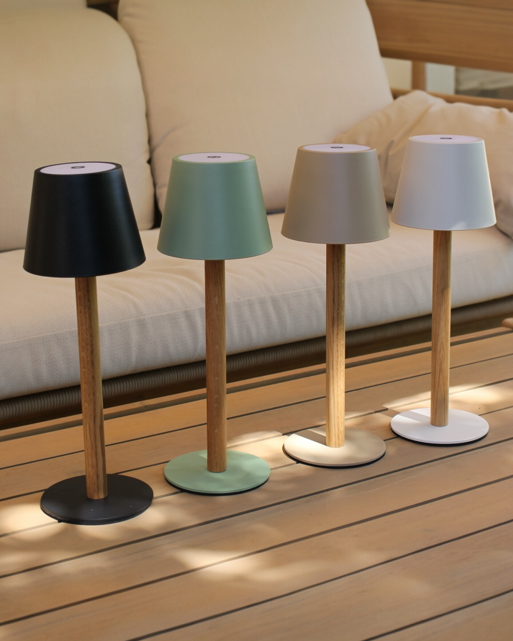 Vier TIMBERCAN LED-Tischlampen in Schwarz, Grün, Beige und Weiß aus Massivholz auf einem Terrassentisch vor einem beigen Outdoor-Sofa