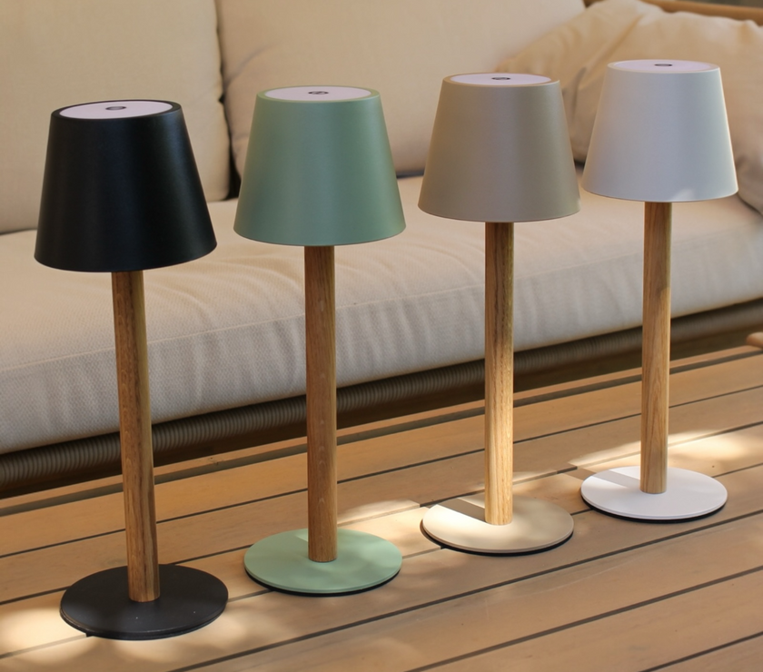 Vier TIMBERCAN LED-Tischlampen in Schwarz, Grün, Beige und Weiß aus Massivholz auf einem Terrassentisch vor einem beigen Outdoor-Sofa