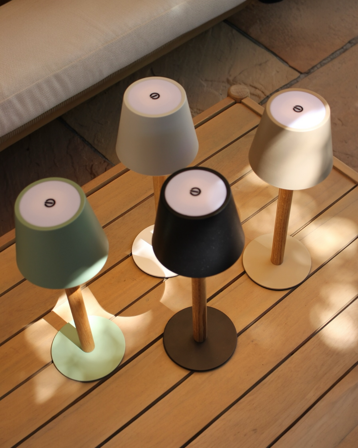 Vier TIMBERCAN Akku-Tischlampen aus Eichenholz in Schwarz, Grün, Beige und Weiß auf Terrassentisch – kabellose LED-Designleuchten für Indoor & Outdoor
