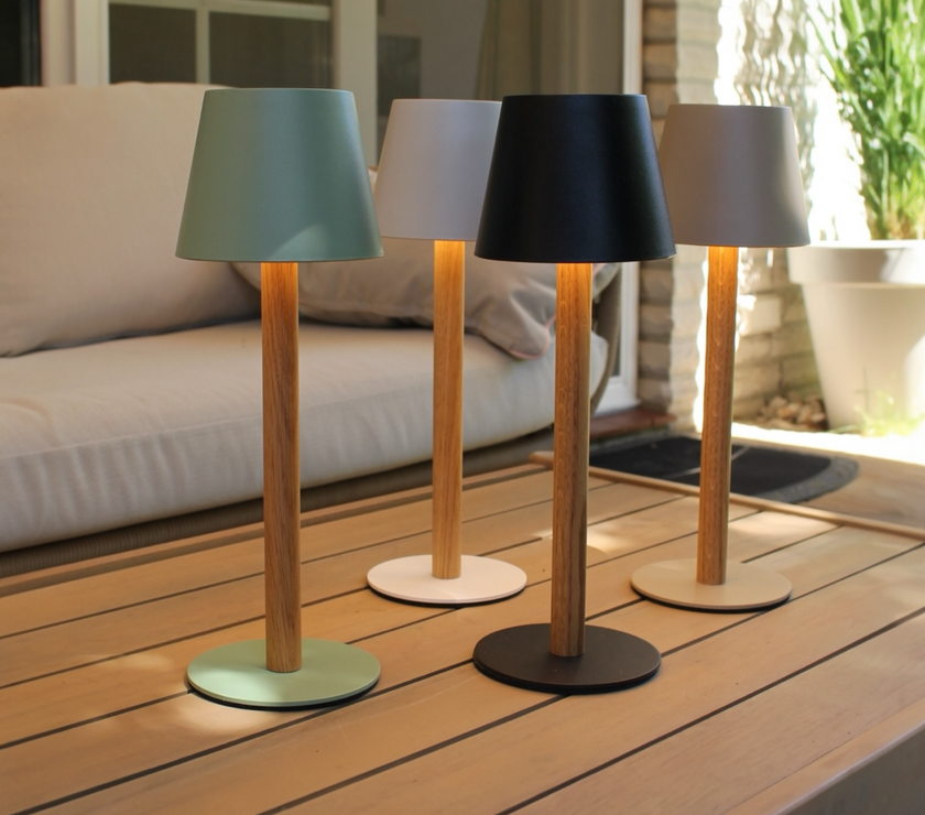 Vier beleuchtete TIMBERCAN Akku-Tischlampen aus massivem Eichenholz in Schwarz, Grün, Beige und Weiß auf einem Terrassentisch – kabellose LED-Designleuchten für Indoor & Outdoor