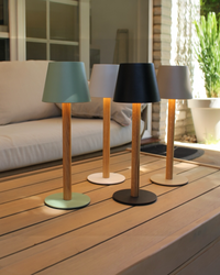 Vier beleuchtete TIMBERCAN Akku-Tischlampen aus massivem Eichenholz in Schwarz, Grün, Beige und Weiß auf einem Terrassentisch – kabellose LED-Designleuchten für Indoor & Outdoor